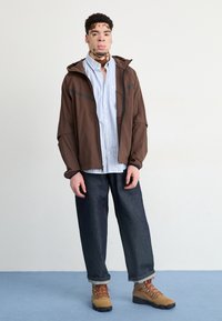 Homme atteint de vitiligo portant une veste marron, une chemise rayée bleu clair, un jean foncé et des bottes de randonnée beige, debout sur un sol bleu contre un mur blanc.