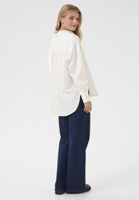 Valge pikkade varrukatega shirt, millel on seotud seljaosa, kombineeritud tumehallide laiade pükstega ja mustade jalanõudega, lihtsa valge taustaga.
