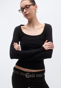 Μαύρο μακρυμάνικο τοπ με φαρδιά στρογγυλή λαιμόκοψη, κατασκευασμένο από ριμπ ύφασμα. Συνδυασμένο με μαύρα τζιν που διαθέτουν ζώνη με καρφιά.