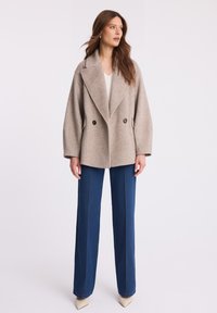 Podwójnie zapinany, jasnobeżowy płaszcz z wełnianej mieszanki z dużym kołnierzem i czarnymi guzikami, w połączeniu z niebieskimi dżinsami o wysokim stanie i beżowymi obcasami.