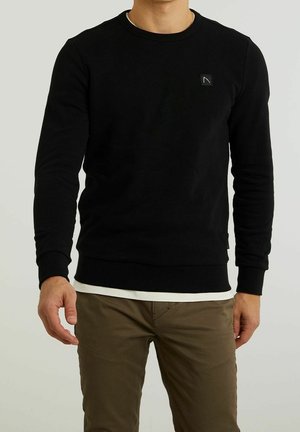 Homme portant un sweat à manches longues noir avec un petit logo sur la poitrine, superposé à une chemise blanche, et un pantalon marron, debout devant un fond uni.