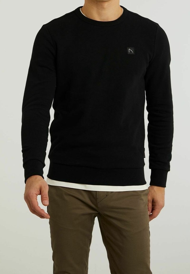 Homme portant un sweat à manches longues noir avec un petit logo sur la poitrine, superposé à une chemise blanche, et un pantalon marron, debout devant un fond uni.