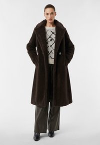 Cappotto in finta pelliccia marrone con ampio colletto, bottoni doppi e tasche laterali, indossato sopra un maglione leggero e pantaloni scuri aderenti.