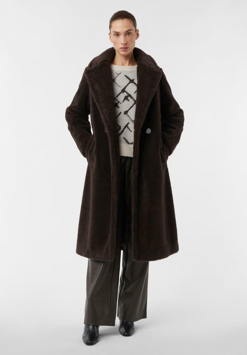 Cappotto in finta pelliccia marrone con ampio colletto, bottoni doppi e tasche laterali, indossato sopra un maglione leggero e pantaloni scuri aderenti.