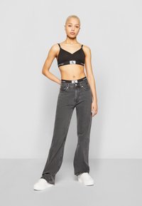 Svart Calvin Klein-bh med logo-stripe og grå vide bukser. Modellen har på seg hvite sneakers. Glatt stoff og rene linjer gjennom hele antrekket.