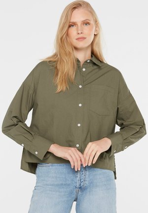 Blonde Frau mit offenem Haar, die ein olivgrünes, geknöpftes Hemd und hellblaue Jeans trägt, stehend vor einem schlichten weißen Hintergrund.