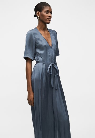 Γυναίκα με σκουρόχρωμο δέρμα και κοντά μαλλιά, που φοράει μεταξωτό μπλε φόρεμα με V λαιμόκοψη, κοντά μανίκια και ζώνη στη μέση, στέκεται μπροστά σε απλό φόντο.