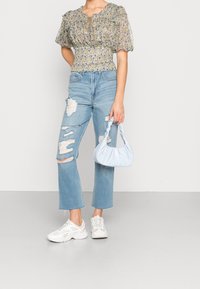 Blus med blommönster och puffärmar, kombinerad med ljusblå slitna jeans och vita sneakers. Håller en ljusblå handväska med vriden handtag.