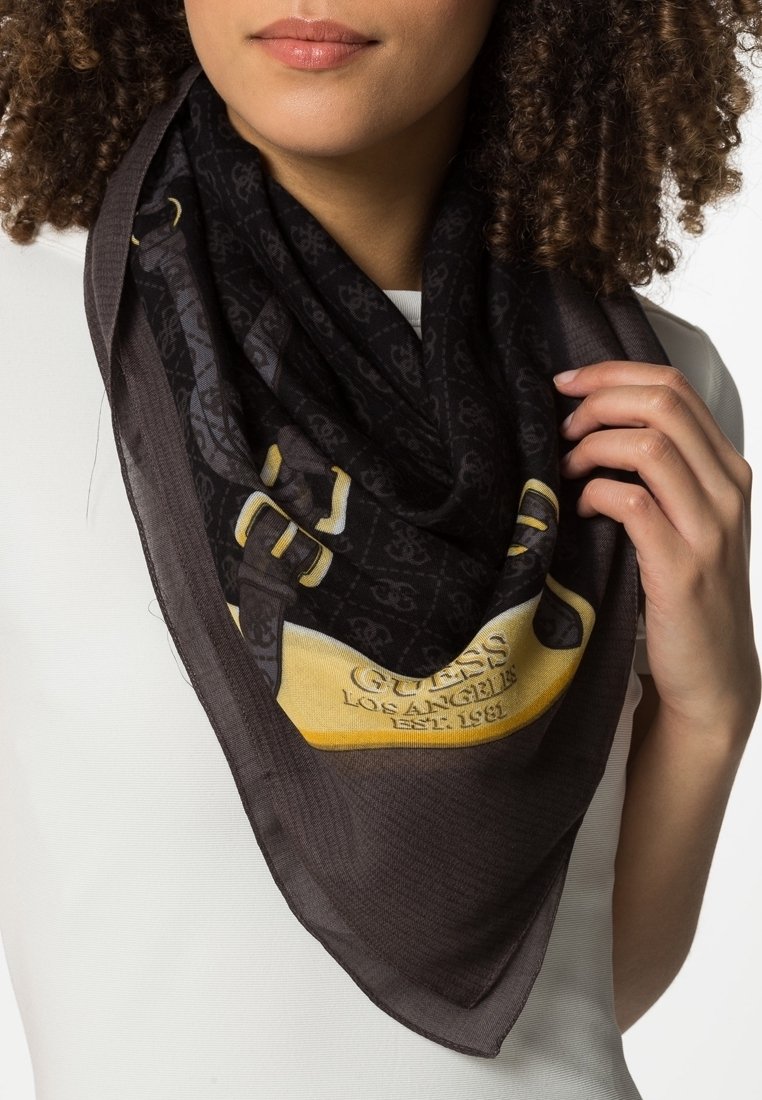 Femme aux cheveux bouclés portant une écharpe sombre à motifs avec une boucle jaune et le texte « GUESS LOS ANGELES EST. 1981 » sur un haut blanc.
