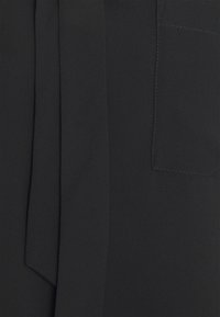 Svart tyg med en slät textur, med en knytdetalj och en ficka. Enkelt design med rena linjer och inga synliga mönster.