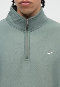 Ketvirčio užtrauktuko džemperis prislopintų žalių audinių. Apatinė dalis turi sidabro užtrauktuką ir baltą Nike logotipą kairėje krūtinės pusėje. Lygi tekstūra, kasdienis dizainas.