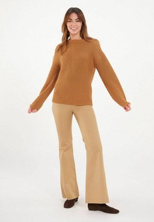 Pull en tricot marron avec un motif en V, manches longues, associé à un pantalon beige évasé et des bottines marron. Texture lisse, coupe décontractée.