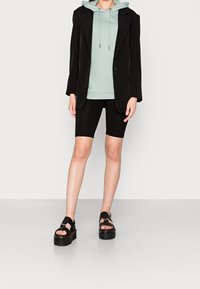 Blazer nero, felpa leggera verde, pantaloni corti neri da ciclista e sandali neri con platform. Tessuto liscio, design a strati e coulisse regolabile sulla felpa.