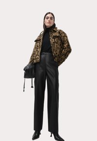 Veste en peluche avec imprimé brun et noir, col roulé noir, pantalon en cuir noir taille haute et chaussures à talons noirs, tenant un sac noir.