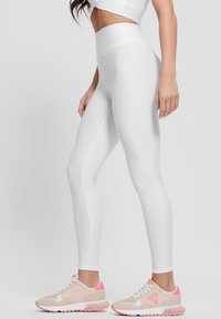 Žena v texturovaných bílých legínách s vysokým pasem a béžovo-růžových teniskách stojící na jednobarevném světlém pozadí.