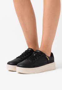 Baskets en cuir noir avec une épaisse semelle couleur crème, dotées de lacets noirs et de détails en accent vert. Marque "Levi's" dorée sur le côté.