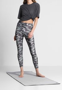 Modelka stoi na szarej macie do jogi, ubrana w zawiązaną czarną bluzkę i legginsy w kamuflaż, przyjęta w zrelaksowanej sportowej pozycji.