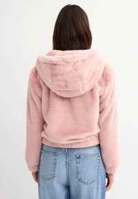 Sudadera de felpa sintética de color rosa claro y suave con capucha. Cuenta con un diseño cropped y dobladillo elástico. Combinada con jeans de mezclilla azul de cintura alta.