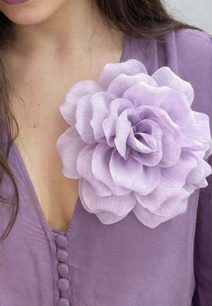 Kobieta w fioletowej bluzce z guzikami pokrytymi tkaniną oraz dużym, jasnofioletowym kwiatem z tkaniny przypiętym blisko dekoltu.