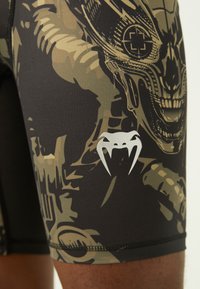 Shorts neri adattati con un design mimetico verde e beige, caratterizzati da una grafica dettagliata e un piccolo logo argentato con accenti a forma di zanna.