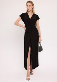 Czarna maxi sukienka z głębokim dekoltem w kształcie litery V, krótkimi rękawami, skręconym detalem z przodu i rozcięciem do uda. Srebrne sandały na obcasie z odkrytymi palcami i teksturowana torebka.