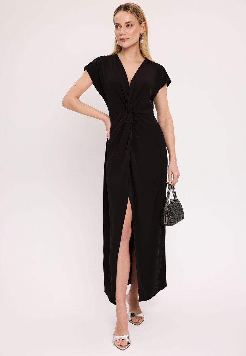 Czarna maxi sukienka z głębokim dekoltem w kształcie litery V, krótkimi rękawami, skręconym detalem z przodu i rozcięciem do uda. Srebrne sandały na obcasie z odkrytymi palcami i teksturowana torebka.