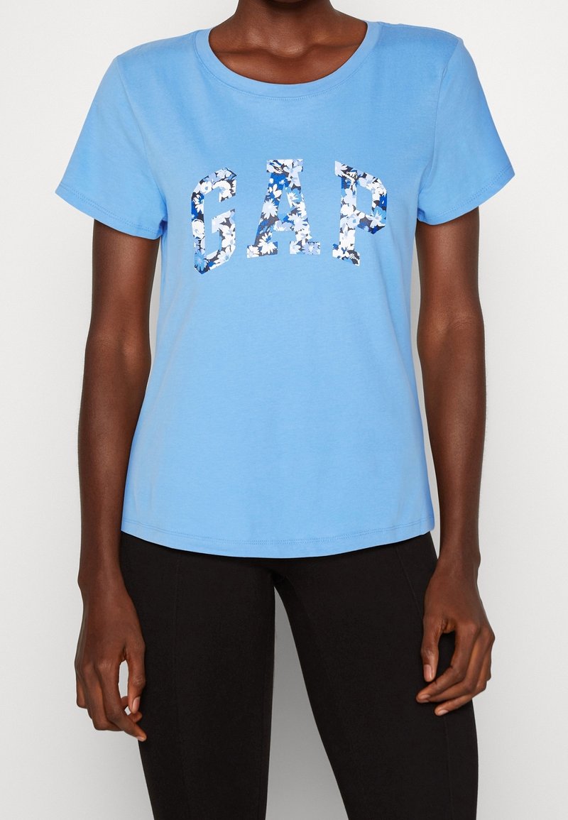 Personne portant une chemise bleu clair à manches courtes avec le texte "GAP" en motif floral bleu et blanc, associée à un pantalon noir.