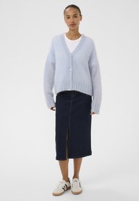 Cardigan en tricot flou bleu clair avec col en V, boutons sur le devant et manches larges ; associé à une jupe en denim foncé avec une fente à l'avant. Tee-shirt blanc en dessous.