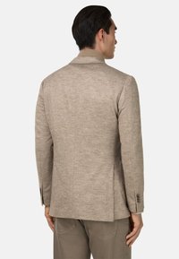 Texturerad beige blazer med ett subtilt hundtandsmönster, slagkrage och knäppta manschetter, kombinerad med avsmalnande bruna byxor.