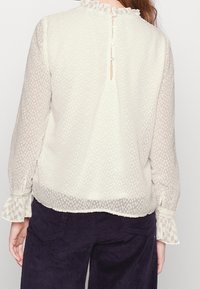 Vaalean kerma väri blouse, jossa on teksturoituja pilkkuja, röyhelökaulus ja -hihat, sekä napitus takaosassa. Läpinäkyvä, virtaava kangas.