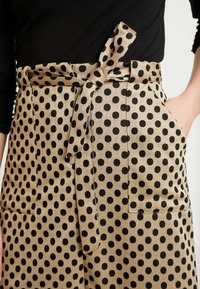 Pantalones beige con estampado de lunares negros, cinturilla atada, y dos bolsillos frontales, con detalles de costura prominentes.