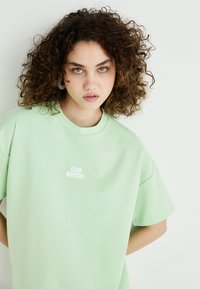 Tyrkysovo-zelené oversize tričko z bavlny, s malým bielym logom "CLUB MATCHA" na prednej strane. Krátke rukávy a voľný strih.