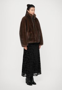 Giacca di pelliccia marrone con colletto alto, chiusura con zip e due tasche, indossata sopra un lungo vestito in pizzo nero, abbinata a stivaletti neri con tacco.
