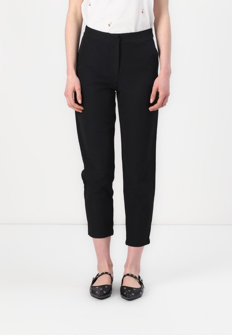 Femme portant un pantalon noir ajusté à la cheville, un haut blanc et des ballerines noires avec des œillets argentés sur fond blanc.