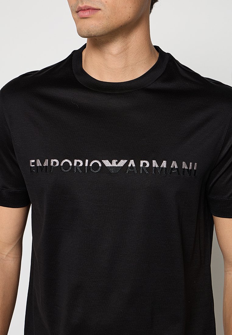 Hombre con una camiseta negra de manga corta con "EMPORIO ARMANI" y el logo del águila Armani en letras metálicas en el pecho.