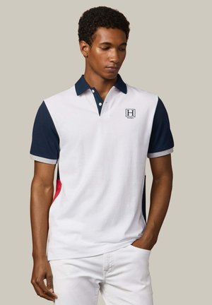 Hombre vistiendo una camiseta tipo polo blanca con mangas y cuello azul marino, pantalones blancos y zapatos tipo mocasín azul marino, de pie contra un fondo beige.