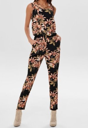 Femme portant une combinaison noire sans manches à fleurs avec des feuilles roses et vertes, des bottines beiges et un collier en or, mains dans les poches.