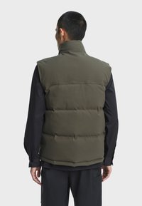 Olivgrüne Steppweste mit hohem Kragen, gesteppte Abschnitte und glatter Oberfläche. Trägt man über einem schwarzen Langarmshirt, mit sichtbaren Nähdetails.