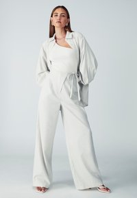 Svijetlosiva oversize košulja s kopčanjem, uparena s hlačama visokog struka širokih nogavica. Rebrasti bijeli top i sandale s remenčićima u bijeloj boji.