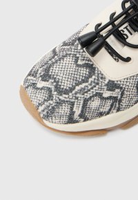 Αθλητικό παπούτσι με υφή από μοτίβο φίδι σε μαύρο και άσπρο, με λαστιχένια σόλα και μαύρα κορδό�νια. Οι λεπτομέρειες περιλαμβάνουν μπεζ επάνω πάνελ.