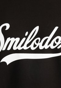 Czarna tkanina z dużym białym napisem "Smilodon" z zakrzywionym podkreśleniem. Materiał wydaje się gładki o matowym wykończeniu.