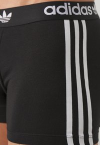 Schwarze Baumwoll-Boxershorts mit einem weißen Adidas-Logo-Bund, die drei vertikale weiße Streifen an den Seiten aufweisen. Glatte Textur.