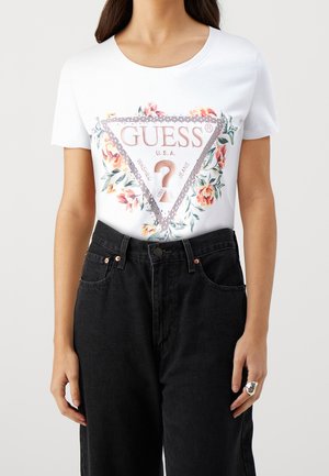 Vrouw draagt een wit T-shirt met een bloemen GUESS driehoekig logo en een zwarte hoge taille jeans, staand met ontspannen armen langs het lichaam.