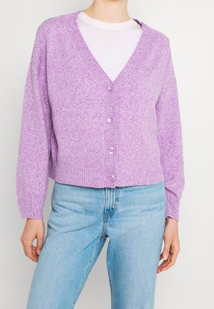 Frau trägt eine lavendelfarbene, geknöpfte Strickjacke über einem weißen Hemd, kombiniert mit hellblauen Jeans, steht vor einem schlichten Hintergrund.