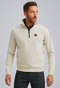 Sudadera color crema con cuello con cremallera, puños y ribete acanalados. Presenta un pequeño parche con logo en el pecho y un bolsillo lateral en la manga.