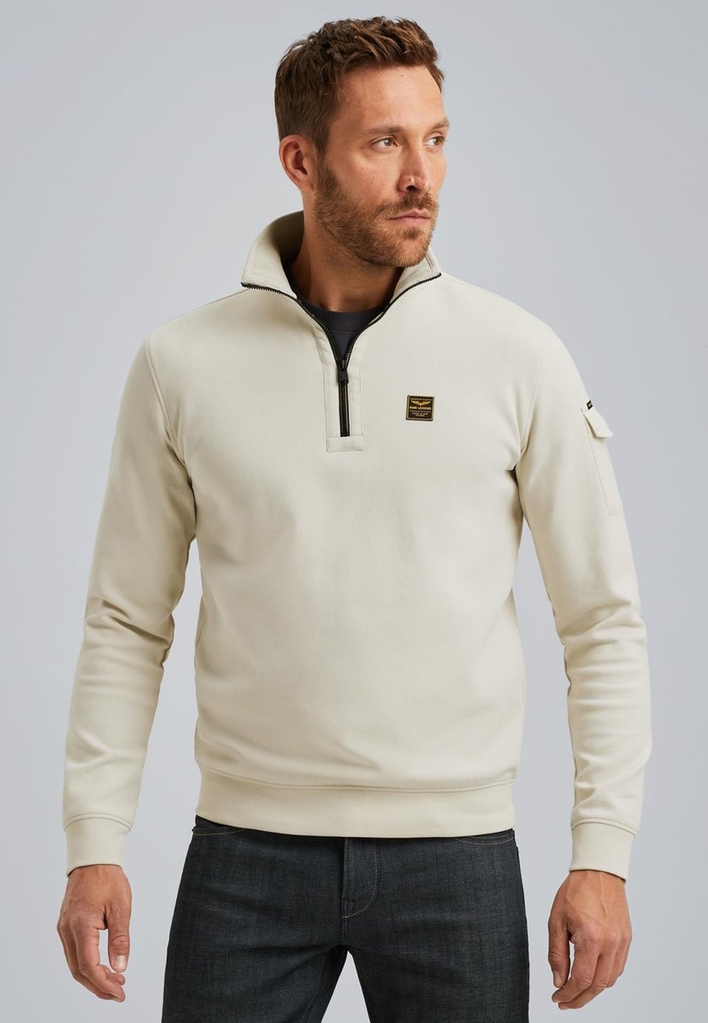 Sudadera color crema con cuello con cremallera, puños y ribete acanalados. Presenta un pequeño parche con logo en el pecho y un bolsillo lateral en la manga.