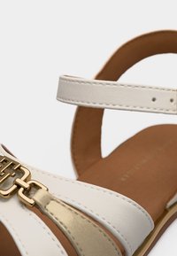 Κοντινή λήψη λευκού και μπεζ σανδαλιού με χρυσή διακοσμητική πόρπη και ρυθμιζόμενο λουράκι στον αστράγαλο πάνω σε ταμπά σόλα.