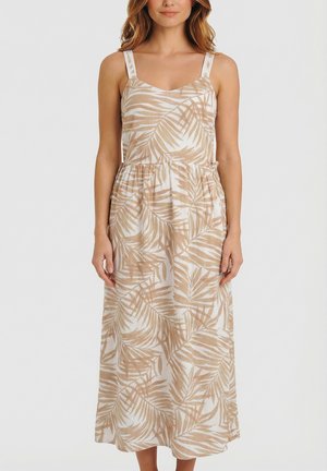 Femme portant une robe longue beige et blanche sans manches avec un motif de feuilles et une taille froncée, debout devant un fond uni.