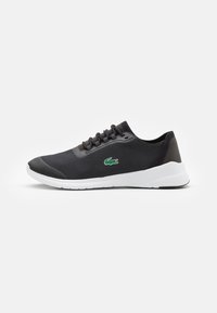 Baskets basses noires avec semelle blanche, lacets noirs, tissu texturé et logo crocodile vert sur le côté, présentées de profil sur fond blanc.