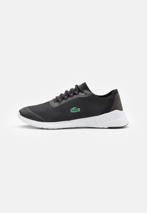 Baskets basses noires avec semelle blanche, lacets noirs, tissu texturé et logo crocodile vert sur le côté, présentées de profil sur fond blanc.